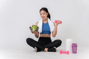 A happy, healthy Asian woman sits on a white studio background, holding a dumbbell and a salad bowl. photo