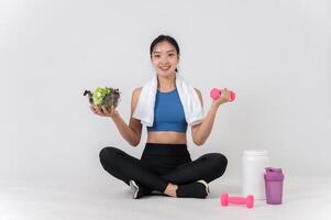 A happy, healthy Asian woman sits on a white studio background, holding a dumbbell and a salad bowl. photo