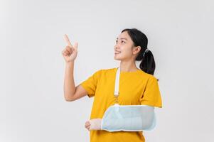 A smiling Asian woman with her broken arm, pointing upward at an empty space, studio background. photo