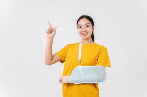 A smiling Asian woman with her broken arm, pointing upward at an empty space, studio background. photo