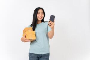 A smiling woman standing against a white studio background, holding cardboard boxes and a smartphone photo