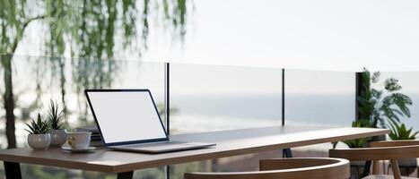 A close-up of a laptop with a white screen mockup on a table on a balcony with a stunning ocean view photo