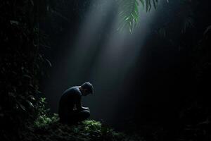 Man kneeling in a dark forest with beams of light breaking through trees photo