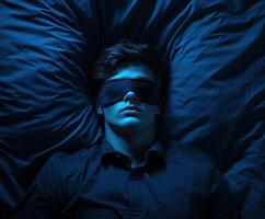 Young man resting in dark bedroom wearing sleep mask and surrounded by blue hues photo