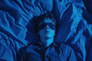 Teenager resting on blue sheets while wearing an eye mask in dim light photo