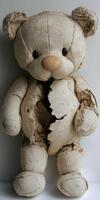 Worn teddy bear with tattered fur and exposed stuffing sits against a wall photo