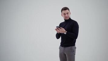 Half-length portrait of man holding a mobile phone. Bearded man in black sweater and grey trousers standing in studio and looking aside with surprise. Slow motion. video