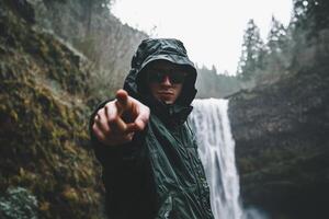 A man in a rain jacket pointing at a waterfall photo