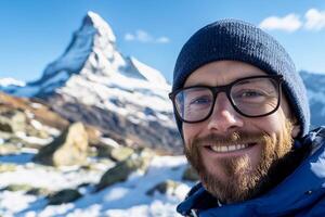 A man with glasses and a blue hat is smiling in front of a mountain photo