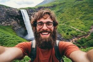 A bearded man with glasses taking a selfie in front of a waterfall photo