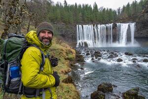 A man with a backpack standing next to a waterfall photo
