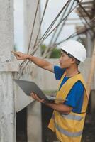 Young handsome asian civil engineer holding tablet looking forward and thinking, planning on construction building background. Evolution construction concept. photo
