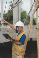 Young handsome asian civil engineer holding tablet looking forward and thinking, planning on construction building background. Evolution construction concept. photo