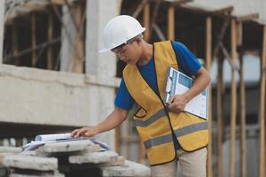 Young handsome asian civil engineer holding tablet looking forward and thinking, planning on construction building background. Evolution construction concept. photo