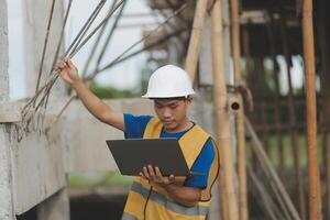 Young handsome asian civil engineer holding tablet looking forward and thinking, planning on construction building background. Evolution construction concept. photo