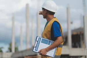 Asian engineer handsome man or architect looking forward holding paperwork blueprint with white safety helmet in construction site. Standing at modern building. photo