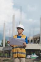 Asian engineer handsome man or architect looking forward holding paperwork blueprint with white safety helmet in construction site. Standing at modern building. photo