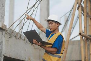 Young handsome asian civil engineer holding tablet looking forward and thinking, planning on construction building background. Evolution construction concept. photo