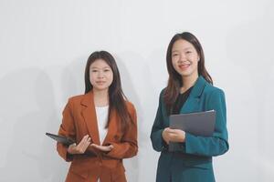 Two Caucasian businesswomen work together using a tablet in a modern office setting, with colleagues engaged in diverse tasks in the background. photo