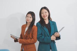 Two Caucasian businesswomen work together using a tablet in a modern office setting, with colleagues engaged in diverse tasks in the background. photo