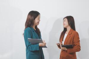 Two Caucasian businesswomen work together using a tablet in a modern office setting, with colleagues engaged in diverse tasks in the background. photo