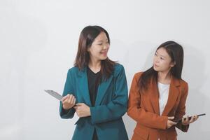 Two Caucasian businesswomen work together using a tablet in a modern office setting, with colleagues engaged in diverse tasks in the background. photo