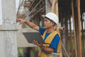 Young handsome asian civil engineer holding tablet looking forward and thinking, planning on construction building background. Evolution construction concept. photo