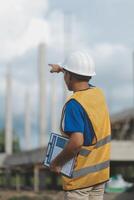 Asian engineer handsome man or architect looking forward holding paperwork blueprint with white safety helmet in construction site. Standing at modern building. photo