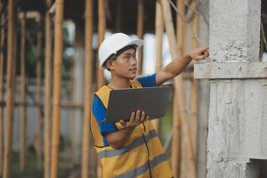 Young handsome asian civil engineer holding tablet looking forward and thinking, planning on construction building background. Evolution construction concept. photo