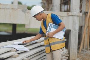Young handsome asian civil engineer holding tablet looking forward and thinking, planning on construction building background. Evolution construction concept. photo
