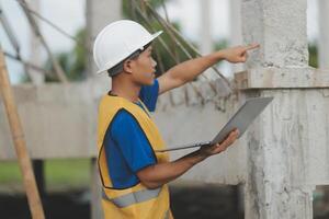 Young handsome asian civil engineer holding tablet looking forward and thinking, planning on construction building background. Evolution construction concept. photo