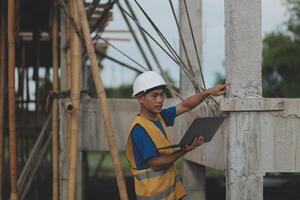 Young handsome asian civil engineer holding tablet looking forward and thinking, planning on construction building background. Evolution construction concept. photo