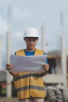 Asian engineer handsome man or architect looking forward holding paperwork blueprint with white safety helmet in construction site. Standing at modern building. photo