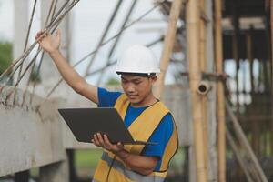 Young handsome asian civil engineer holding tablet looking forward and thinking, planning on construction building background. Evolution construction concept. photo