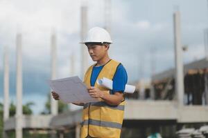 Asian engineer handsome man or architect looking forward holding paperwork blueprint with white safety helmet in construction site. Standing at modern building. photo