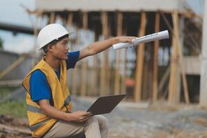 Asian man architect holds blueprint with skyscraper background, Asian male architect builder engineer design working planning confident outdoors in construction site. photo