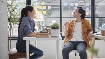 Two Asian workers sitting in chairs at working desks, collaborating and talking at daylight windows in casual small business office, online business job coworkers, positive relationship colleagues. photo