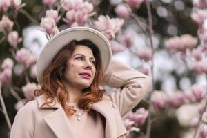 Woman magnolia flowers, surrounded by blossoming trees., hair down, white hat, wearing a light coat. Captured during spring, showcasing natural beauty and seasonal change. photo