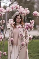 Woman magnolia flowers, surrounded by blossoming trees., hair down, white hat, wearing a light coat. Captured during spring, showcasing natural beauty and seasonal change. photo