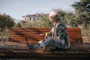 A woman sits on a bench with a laptop open in front of her. She has a cup of coffee in her hand and her jeans are ripped. The scene suggests a relaxed and casual atmosphere. photo