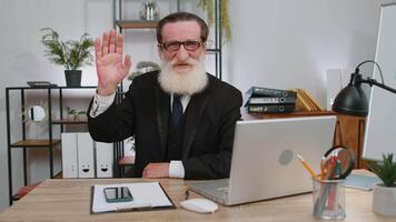 Businessman working on laptop smiling friendly at camera and waving hands gesturing hello at office video