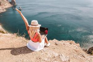 A woman is sitting on a rocky beach with a laptop in front of her. She is wearing a pink top and a straw hat. She is waving her hand in the air, possibly to celebrate or to show excitement. photo