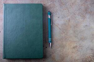 A green bound notebook on a light brown, textured surface, accompanied by a pen photo