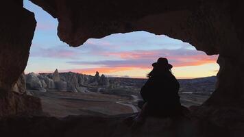A view from the back of a girl in a hat sits on a mountain and admires the beautiful view and dawn or sunset in Cappadocia in Turkey. Goreme red valley fairy chimneys. Digital detox and soul search video