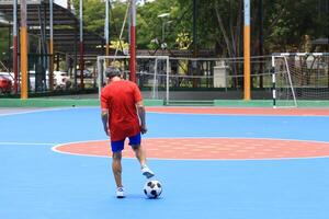 Back view of senior Asian man in retirement age playing soccer football at outdoor court for healthy and fun activity to exercise and workout against aging illness photo