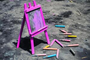 an easel painted in bright baby pink holds a canvas featuring a charcoal drawing accented with soft pastel colors. the ground is scattered with charcoal sticks and chalks photo
