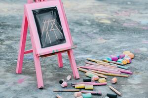 an easel painted in bright baby pink holds a canvas featuring a charcoal drawing accented with soft pastel colors. the ground is scattered with charcoal sticks and chalks photo