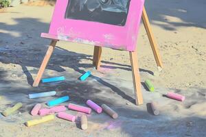 an easel painted in bright baby pink holds a canvas featuring a charcoal drawing accented with soft pastel colors. the ground is scattered with charcoal sticks and chalks photo
