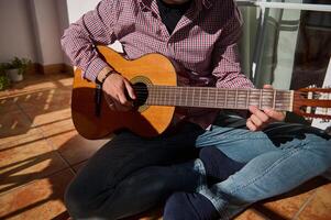 Man playing acoustic guitar while sitting on a sunlit tiled floor photo