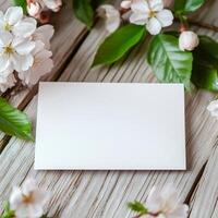 Blank card surrounded by delicate pink flowers and green leaves on rustic wooden background photo